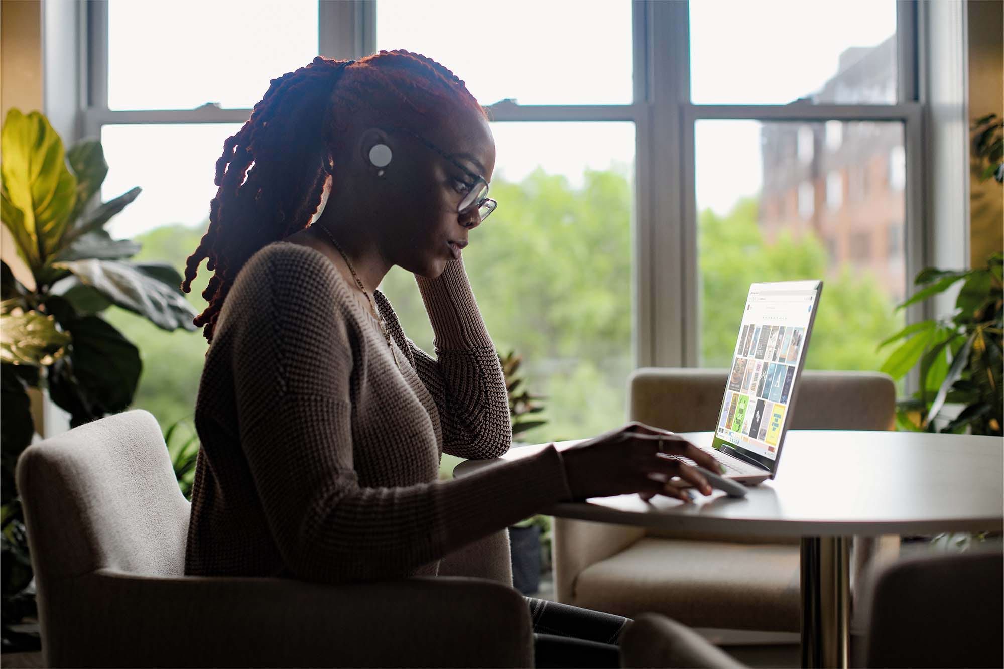 A woman with dreadlocks, wearing glasses and a sweater, is focused on her laptop