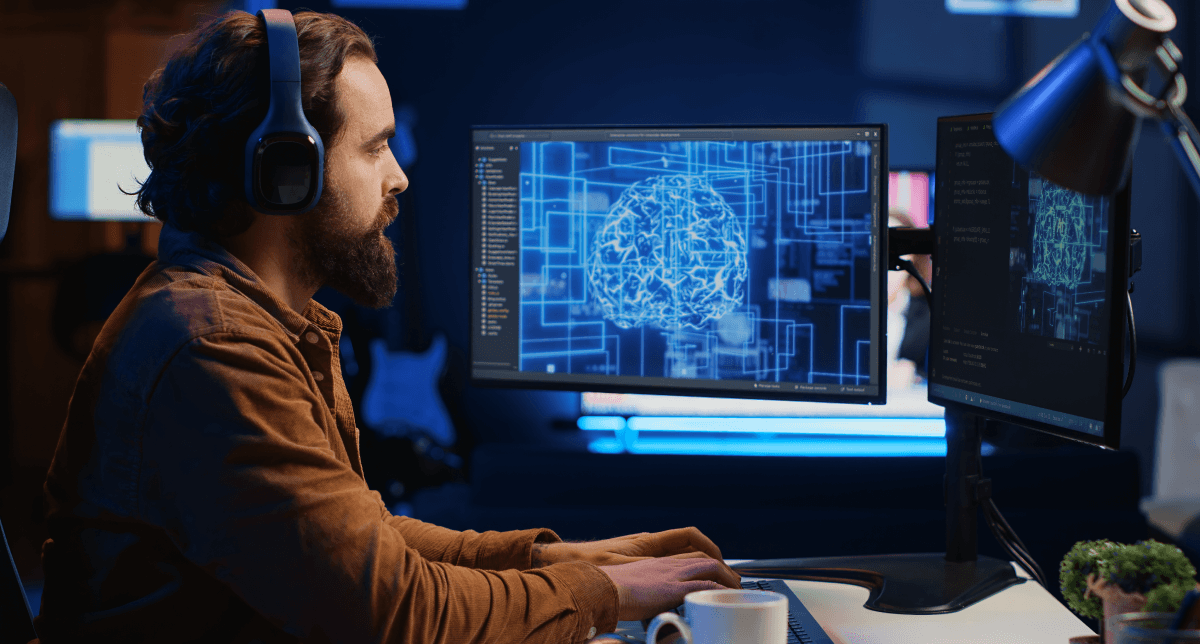 A man with a beard and headphones is focused on two computer screens