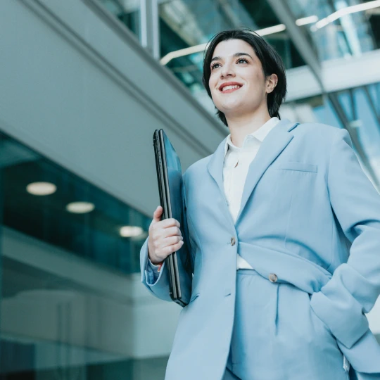 A smiling woman in a blue suit, holding a laptop, stands in a modern office building