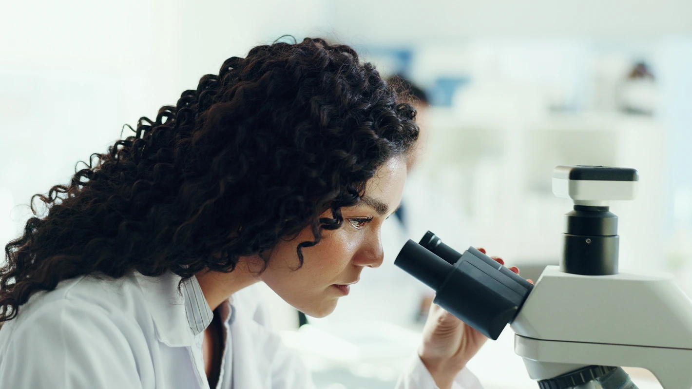 A curly-haired woman in a white lab coat intently looks through a microscope in a brightly lit lab setting