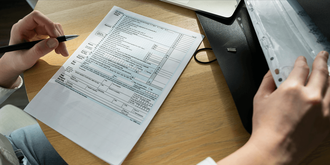 Person holding a pen while filling out a tax form on a light-colored wooden desk