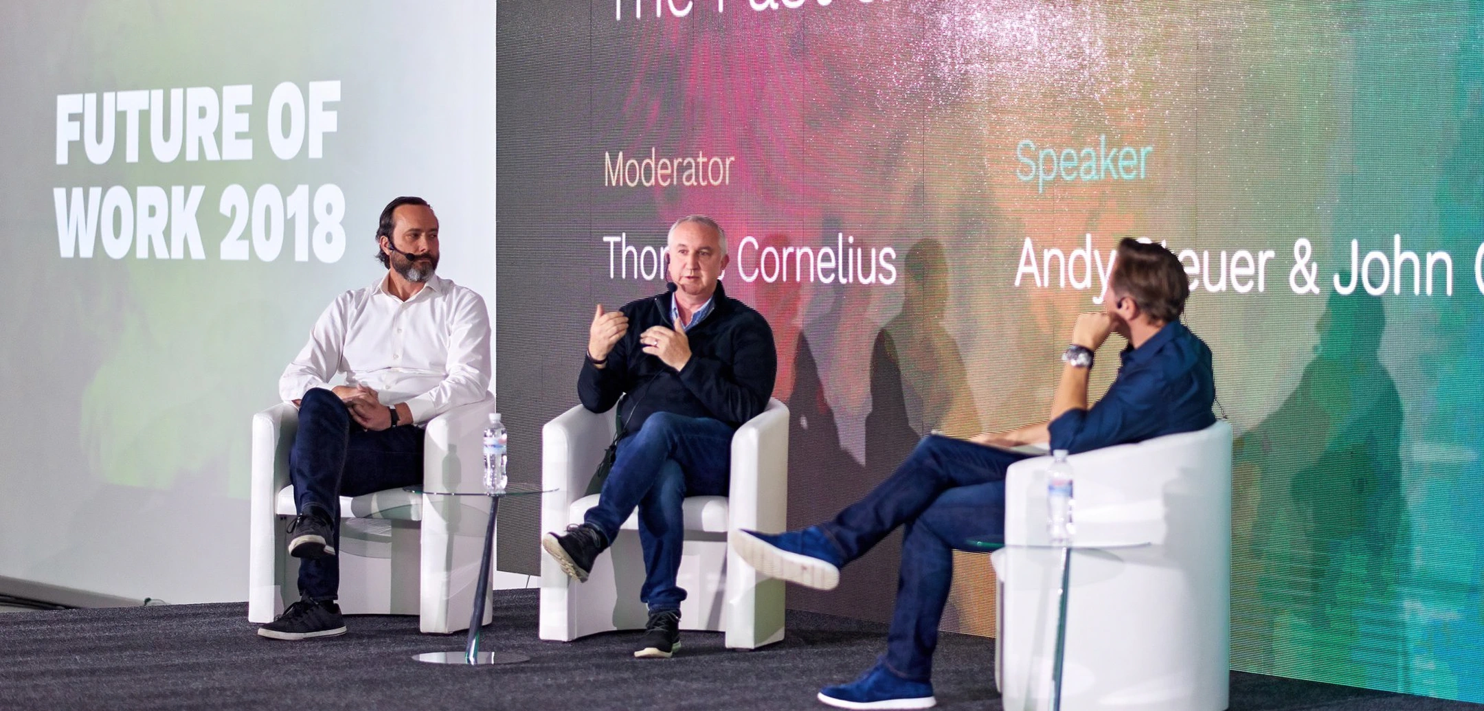 Three men in headsets are sitting on white chairs on the stage
