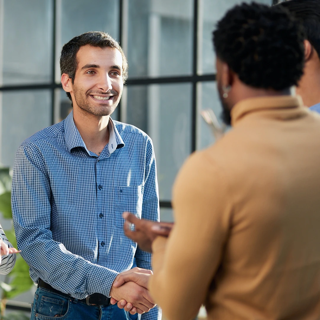 Smiling man in a blue checkered shirt shakes hands with another man in a tan sweater, in a modern office setting