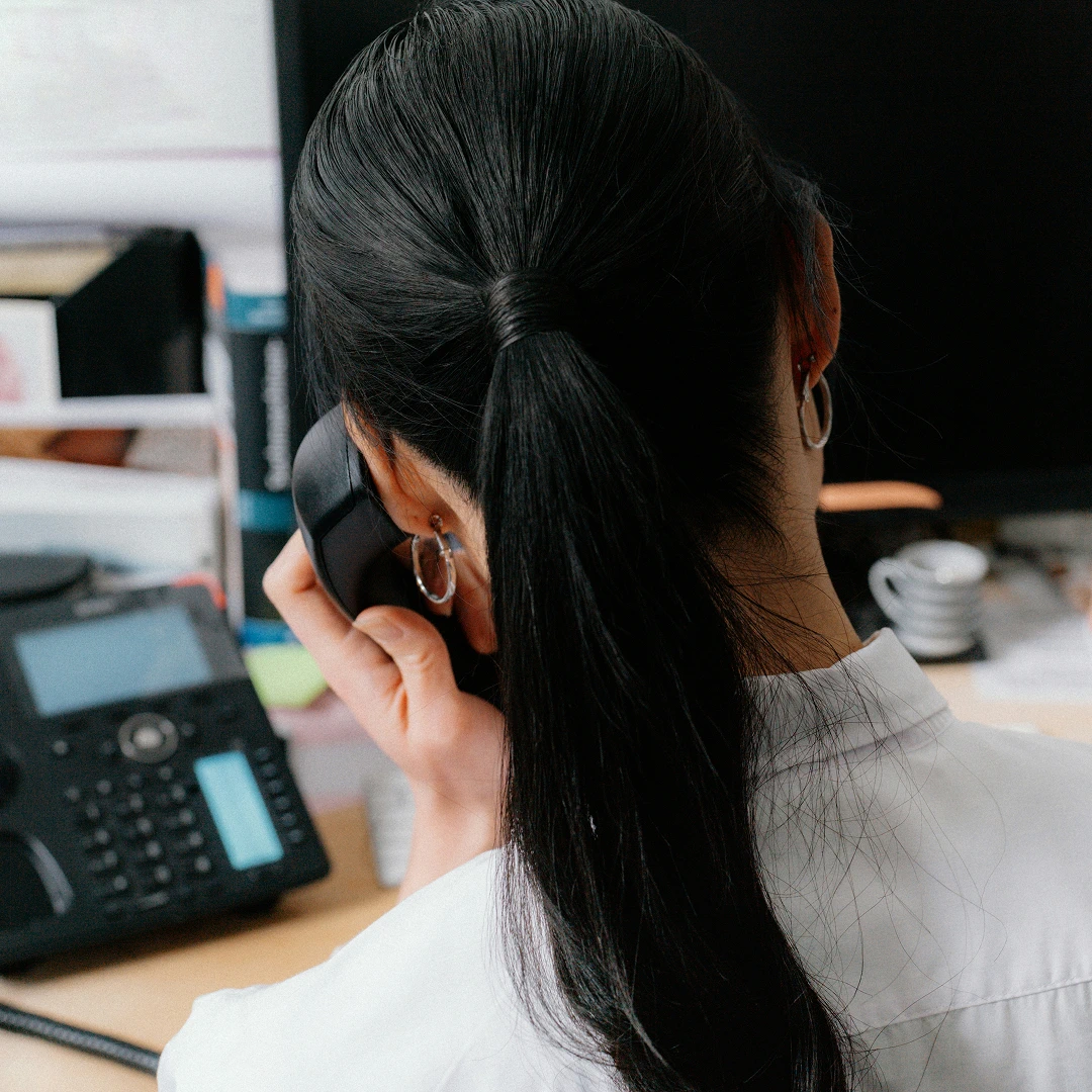 View from behind of a woman with dark hair in a ponytail talking on a black landline phone, wearing a white shirt and hoop earrings