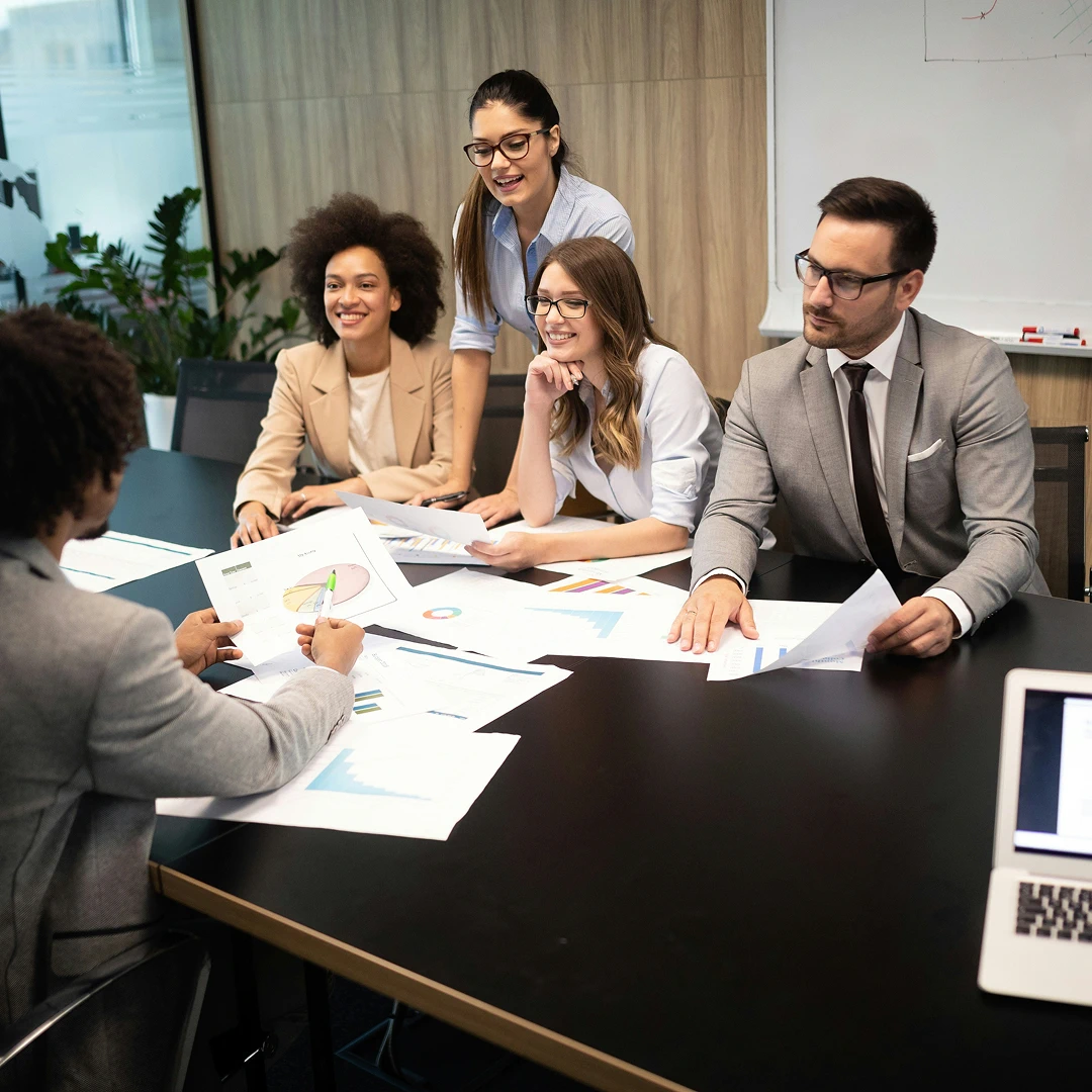 Business professionals, including three women and two men, meeting around a large table covered with documents, charts, and graphs