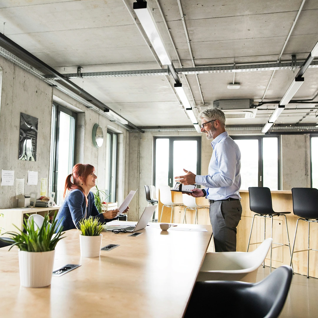 Modern office space with a long table, featuring two colleagues discussing paperwork