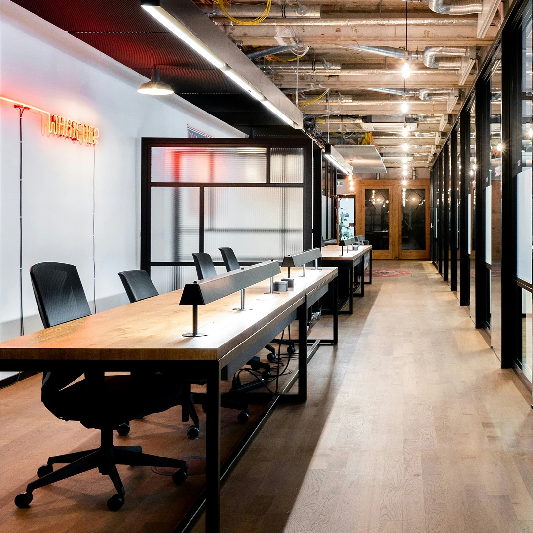 Modern, open-plan office space with long wooden desks, black chairs, and an exposed ceiling