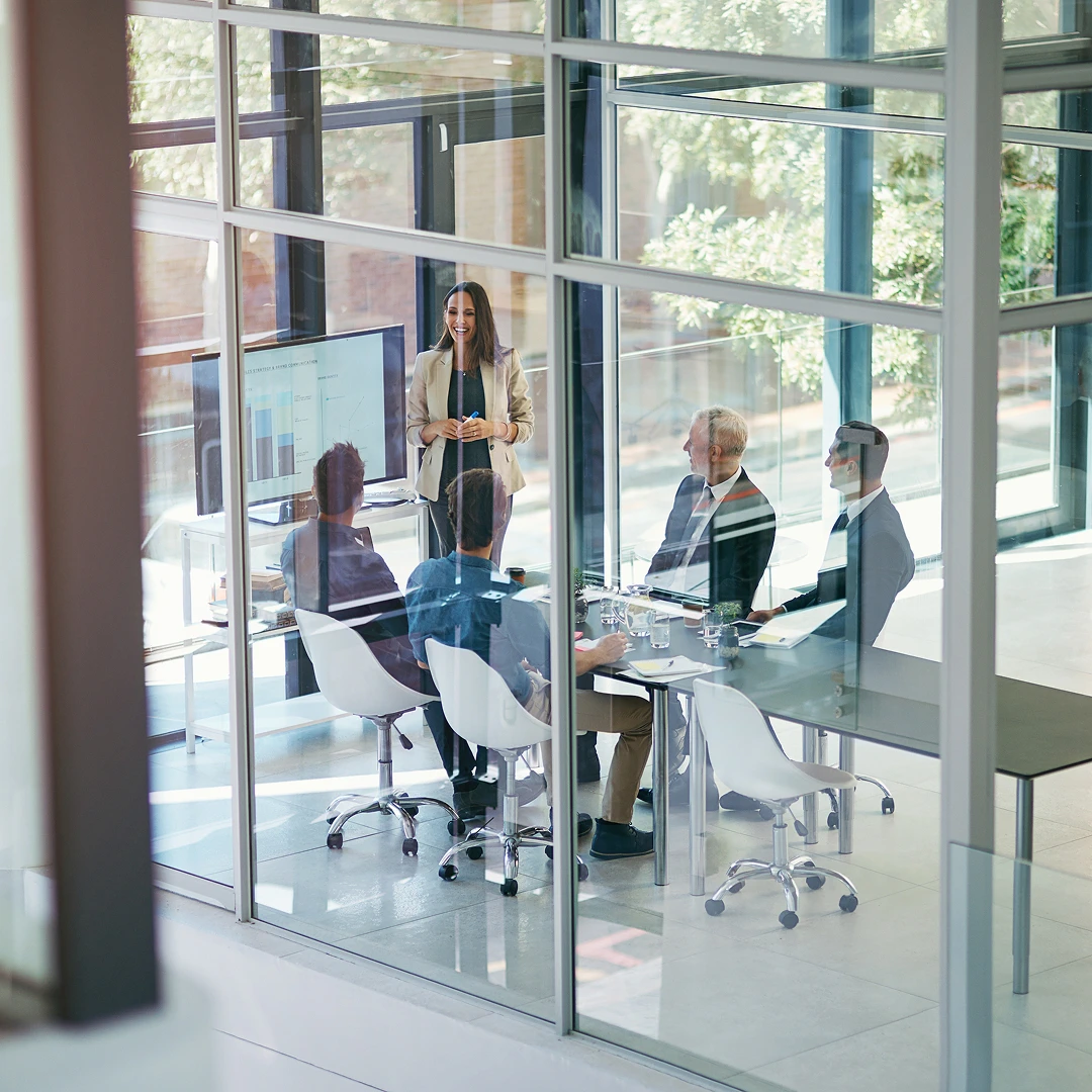 A business meeting in a conference room with glass walls. A woman is giving a presentation, showcasing charts and graphs.