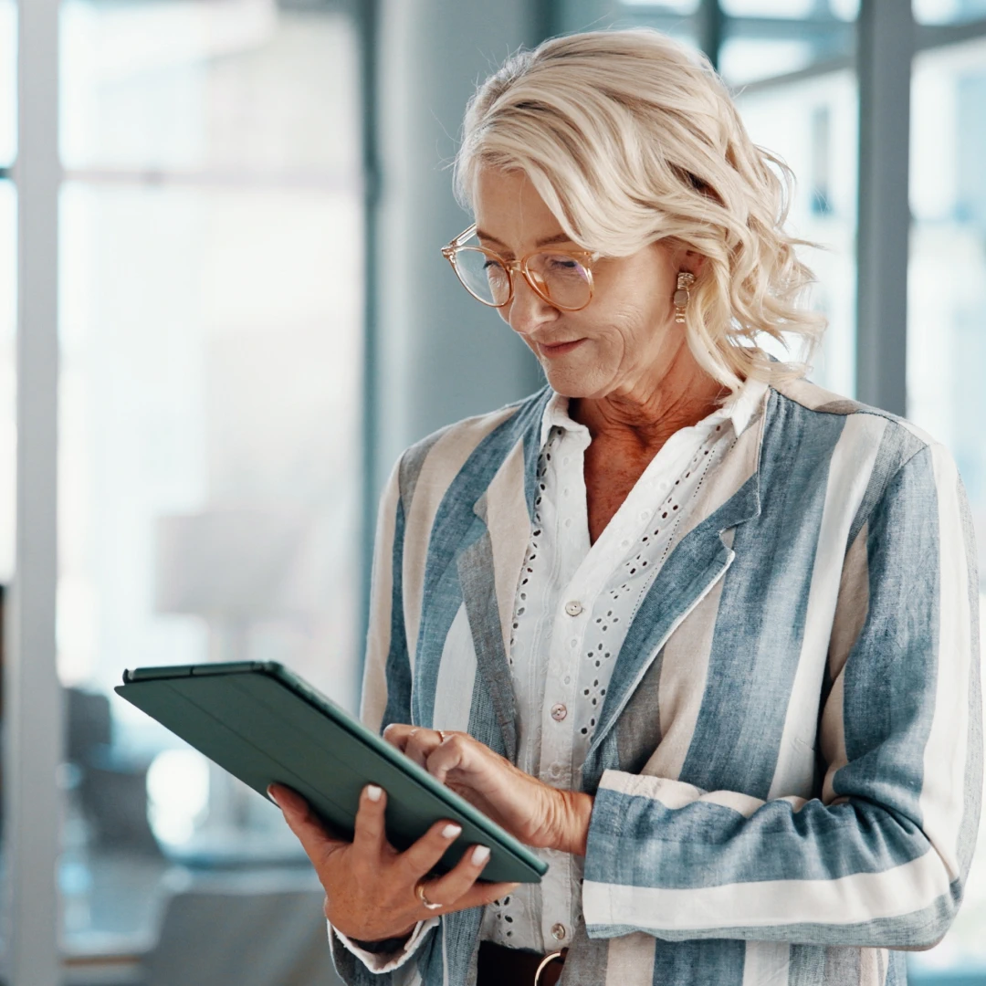 Mature woman with blonde hair and glasses is using a tablet. She is wearing a striped blazer and a white blouse
