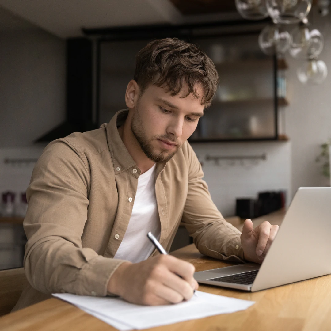 Man in a brown shirt using a laptop and writing on a paper on a wooden table.