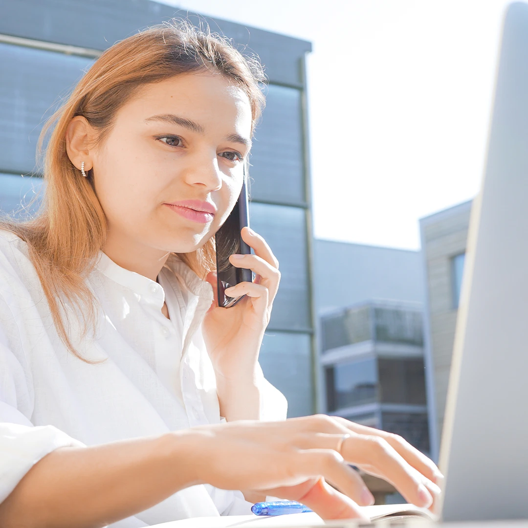 Young woman with light brown hair in a white shirt, using a laptop and talking on her smartphone