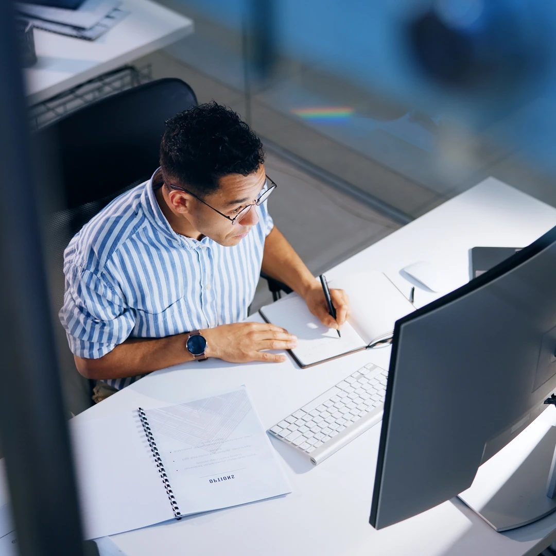 Man with short hair, wearing a striped shirt and glasses, is working at a white desk while looking at a computer monitor and taking notes in a notebook with a pen