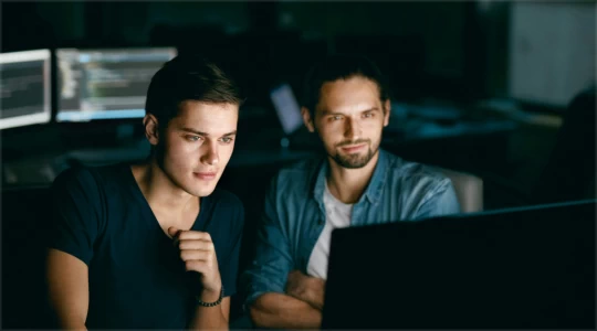 two guys looking at a screen, the bearded one smiling